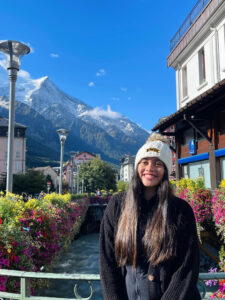 Female student with long hair standing in front of a mountainscape in France wearing a UMBC beanie.